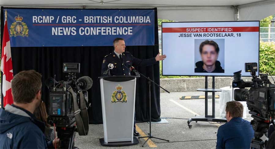 RCMP officer standing at a podium during a press conference, gesturing towards a large screen displaying the text "SUSPECT IDENTIFIED: JESSE VAN ROOTSELAAR, 18" alongside a blurred photo of a young person. Professional video cameras are visible in the foreground filming the announcement.