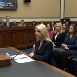 Attorney General Pam Bondi sits sternly at a witness table in a crowded House hearing room, looking straight ahead, while a row of solemn women—survivors of Jeffrey Epstein—sit directly behind her in the public gallery.